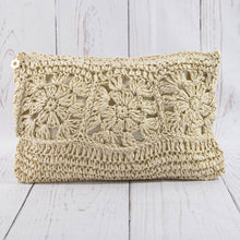 Load image into Gallery viewer, A handmade crochet clutch bag in a natural beige straw color, featuring intricate floral granny square patterns, displayed on a white-washed wooden plank surface.
