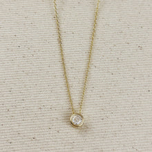 Load image into Gallery viewer, A delicate yellow gold chain necklace with a small bezel-set round clear gemstone pendant, displayed on a textured beige fabric background.