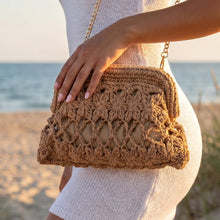 Load image into Gallery viewer, A khaki crocheted shoulder bag with a floral pattern, textured stitching, and a gold-tone chain strap, shown against a beach background.
