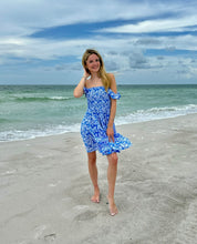 Load image into Gallery viewer, A woman stands on a sandy beach wearing a blue and white floral off-the-shoulder mini dress, with the ocean and cloudy sky in the background.