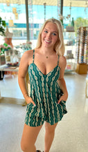 Load image into Gallery viewer, A smiling woman poses indoors wearing a green patterned sleeveless romper with a V-neckline, thin straps, and pockets. She is standing in a brightly lit boutique.