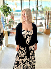 Load image into Gallery viewer, A woman with blonde hair stands inside a boutique shop wearing a black floral midi dress with a black cardigan. The shop has large windows in the background showing a sunny outdoor scene.