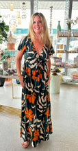 Load image into Gallery viewer, A woman modeling a dark green maxi dress featuring a bold floral print in orange and cream. The dress has a V-neckline with a gathered tie-front detail, short flutter sleeves, and a relaxed, flowy silhouette. The background is a bright boutique.