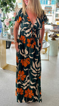 Load image into Gallery viewer, A close-up image of a woman wearing a dark green maxi dress with a bold orange and cream floral print. The background is of a bright boutique.