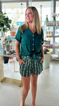 Load image into Gallery viewer, A woman modeling a dark green button up tie front top, paired with dark green shorts with a cream geometric pattern, with a background of a bright boutique.