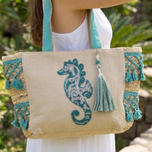 Load image into Gallery viewer, Beige tote bag with teal embroidery seahorse and tassel, held by a person in a white dress outdoors.