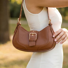 Load image into Gallery viewer, A brown shoulder bag with a curved silhouette, decorative stitching, and a wide center strap with a gold-tone buckle, worn against a white dress in a bright outdoor background.