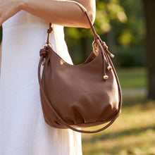 Load image into Gallery viewer, A brown leather shoulder bag with a slouchy crescent shape, draped over the shoulder of a person wearing a white dress, set against a blurred outdoor background.