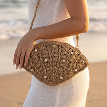 Load image into Gallery viewer, Person holding a woven handbag with pearl embellishments on a beach.
