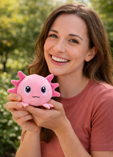 Load image into Gallery viewer, A smiling woman in a dusty rose t-shirt holds a cute pink plush sea urchin toy with large cartoon eyes and small spiky fins, set against a soft-focus green outdoor background.

