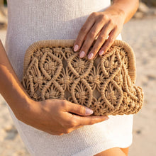 Load image into Gallery viewer, Woven macrame clutch bag with an intricate floral pattern and textured rope detailing, shown against a white dress with a beach background.
