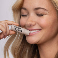 Load image into Gallery viewer, A close-up lifestyle shot of a woman holding a beige Island Coconut lip balm tube to her lips. The tube is labeled 'poppy &amp; pout' in black text.
