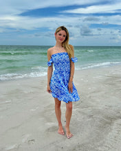 Load image into Gallery viewer, A woman stands on a sandy beach wearing a blue off-the-shoulder floral mini dress, with the ocean and cloudy sky in the background.
