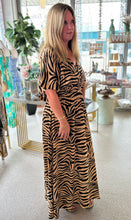 Load image into Gallery viewer, A woman standing in a boutique models a tan and black zebra print maxi dress, and she's facing the side.
