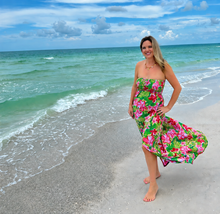 Load image into Gallery viewer, A woman stands on a sandy beach wearing a strapless maxi dress with a bright pink and green tropical floral print. The ocean and blue sky with clouds are visible in the background.
