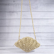 Load image into Gallery viewer, A natural beige woven straw shoulder bag with a scalloped, lace-like openwork pattern and small pearl embellishments, hanging against a light gray wood plank background.
