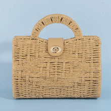 Load image into Gallery viewer, A woven straw handbag with a structured rectangular shape, rounded handles, and a gold-tone clasp, shown against a light blue background.
