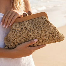 Load image into Gallery viewer, A tan crocheted clutch with an intricate open-knit pattern and a wooden frame closure with round wooden knobs, held against a beach setting.
